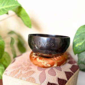 Może przedstawiać: Ciemnoszara miska śpiewająca z wytłoczonymi symbolami spoczywa na pomarańczowo-złotej poduszce, umieszczonej na wzorzystym stołku. Zielone, liściaste rośliny otaczają misę, tworząc spokojne, naturalne otoczenie. Misa jest narzędziem do medytacji.