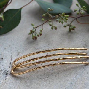 May include: Three gold hair pins with a hammered texture. The pins are curved and have a simple design.