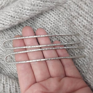 May include: Three silver hair pins with a hammered texture. The pins are held in a hand against a gray knitted background.