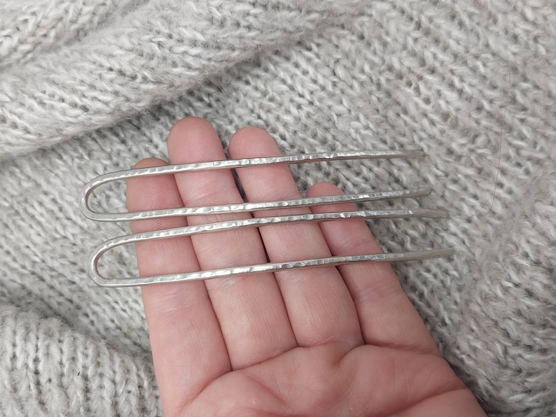 May include: Three silver hair pins with a hammered texture. The pins are held in a hand against a gray knitted background.