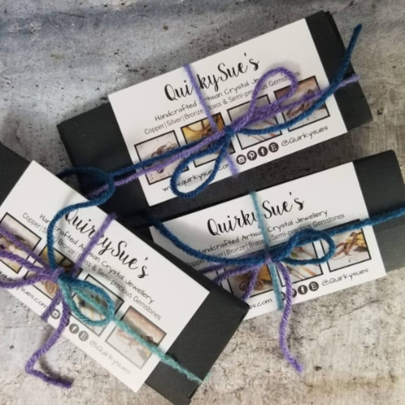 May include: Three black boxes with white labels tied with purple and blue string. The labels read "Quirky Sue's Handcrafted Artisan Crystal Jewellery Copper | Silver | Bronze | Brass & Semi-precious Gemstones" and include a website address and social media icons.