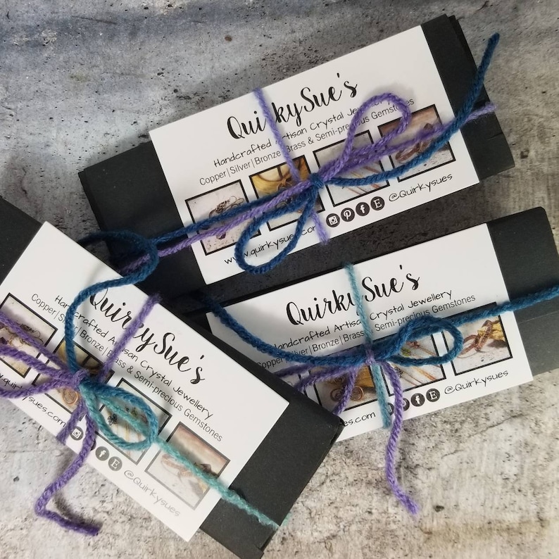 May include: Three black boxes with a purple and blue string tied around them. Each box has a white label with the text "Quirky Sue's" and a description of handcrafted artisan crystal jewelry made with copper, silver, bronze, brass, and semi-precious gemstones. The boxes are stacked on top of each other.