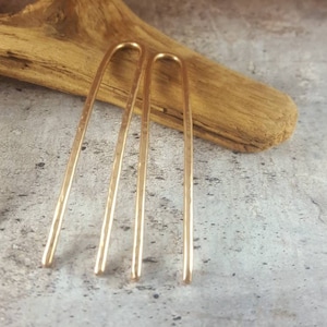 May include: Two gold hair pins with a hammered texture. The pins are long and straight with a slight curve at the top.