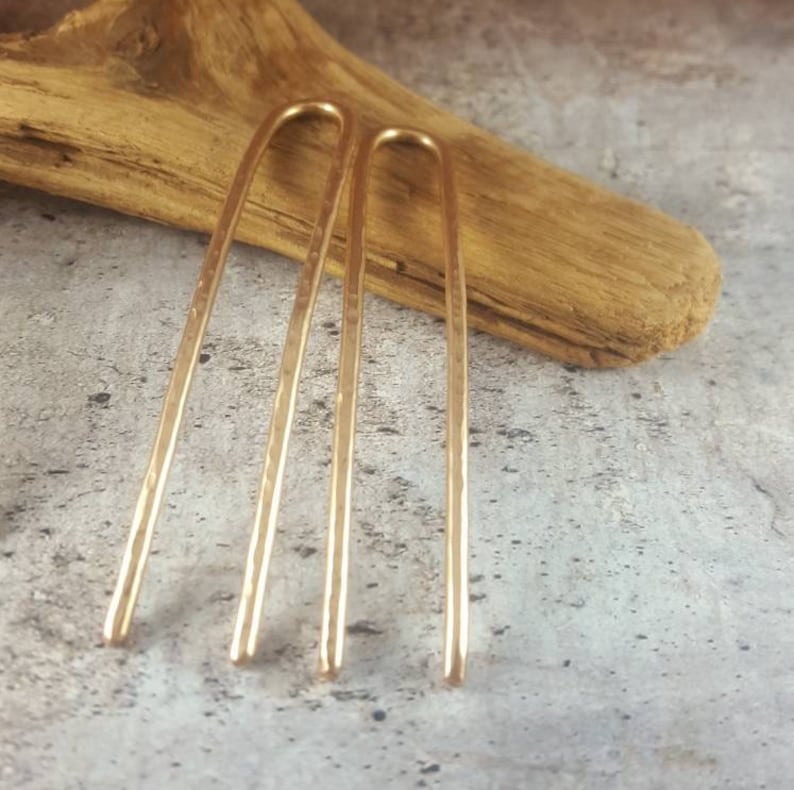 May include: Two gold hair pins with a hammered texture. The pins are long and straight with a slight curve at the top.