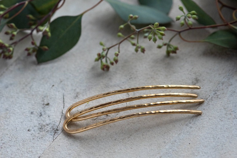 May include: Three gold hair pins with a hammered texture. The pins are curved and have a simple design.