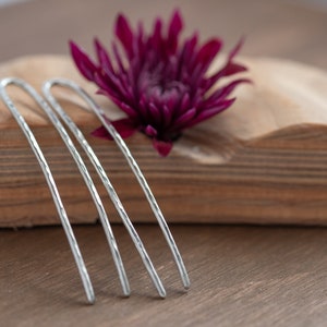 May include: Four silver hair pins with a textured surface. The pins are arranged in a row on a wooden surface.