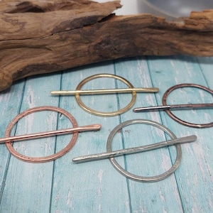 May include: Four metal hair pins in gold, copper, silver, and bronze tones. Each pin features a circular design with a straight bar across the centre. The pins are displayed on a light blue wooden surface.