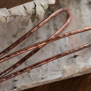 May include: Three hammered copper hair pins, each with a rounded end, are arranged on a piece of birch bark.
