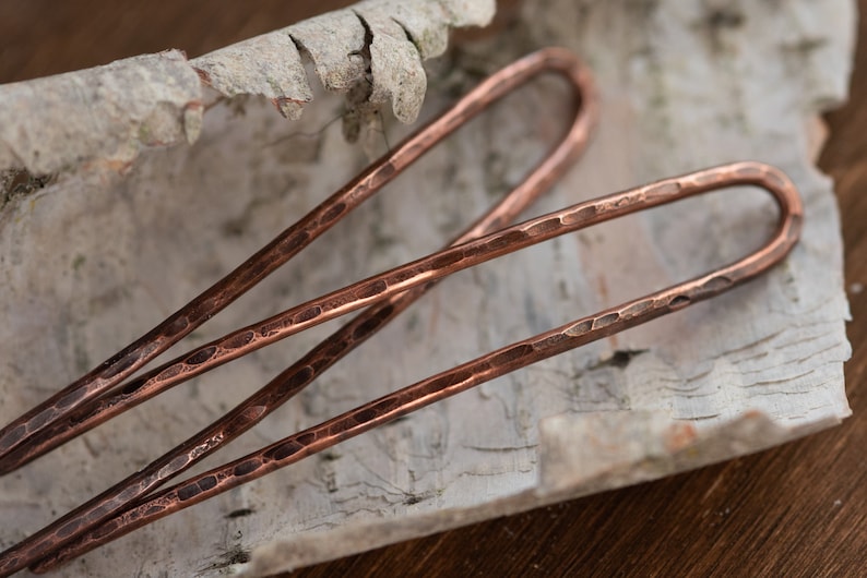 May include: Three hammered copper hair pins, each with a rounded end, are arranged on a piece of birch bark.