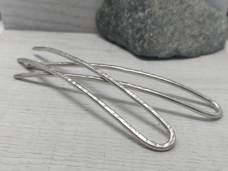 May include: Two silver hair pins with a hammered texture. The pins are curved and have a smooth, rounded end.