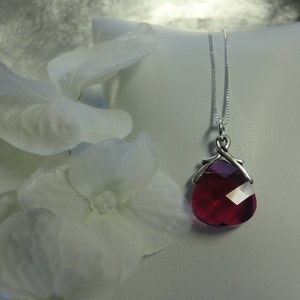 May include: A silver necklace with a faceted, teardrop-shaped, ruby-red pendant. The pendant is suspended from a delicate chain and is displayed on a white surface, with white flowers in the foreground.