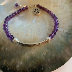 May include: A purple amethyst beaded bracelet with a silver paw print charm and two silver beads. The bracelet is on a white and green shell.