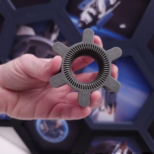 May include: A gray plastic gear with a ring in the center and six spokes. The gear has a textured surface.