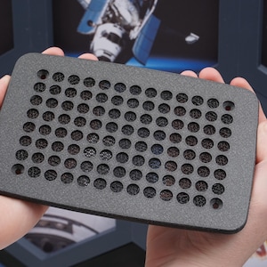 May include: A dark grey rectangular object with a grid of small, evenly spaced holes. The object is held in two hands. The background features images of space and satellites.