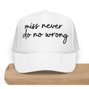 Op de afbeelding: Witte trucker cap met zwarte tekst die "miss never do no wrong" zegt.