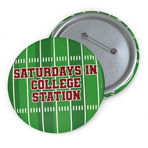 Może przedstawiać: Okrągła przypinka z grafiką zielonego boiska do futbolu amerykańskiego z białymi liniami i napisem SATURDAYS IN COLLEGE STATION pogrubionymi czerwonymi literami z białą obwódką. Z tyłu znajduje się srebrny metalowy mechanizm zapięcia na agrafkę.