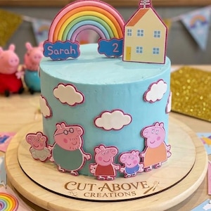 Könnte beinhalten: Ein hellblau glasierter Kuchen, verziert mit Peppa Wutz-Figuren, weißen Wolken, einem Regenbogen-Topper mit dem Namen Sarah und der Zahl 2 sowie einem Haus-Topper. Der Kuchen steht auf einem Holzbrett mit der Aufschrift Cut Above Creations.