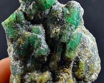 May include: A cluster of emerald crystals in a rough matrix. The crystals are a vibrant green color and have a glassy luster. The matrix is a dark gray color and has a rough texture.