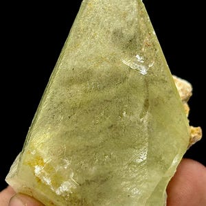 May include: A translucent, light green mineral specimen with a crystalline structure. The stone has a triangular shape with a pointed tip and a rough, textured surface. The color is a pale, yellowish-green with some darker green inclusions.