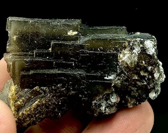 Puede incluir: Un espécimen mineral oscuro y estratificado con una estructura cristalina. El mineral tiene un color profundo, casi negro, con toques de verde y marrón. La superficie tiene un aspecto rugoso y texturizado, con algunas zonas brillantes.