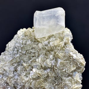 May include: A clear, rectangular crystal sits atop a bed of silvery-grey mica. The crystal is translucent and reflects light.