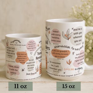 May include: Two white ceramic mugs with inspirational quotes and floral accents. The mugs, labeled 11 oz and 15 oz, feature quotes in English with a light beige and pink color palette. The mugs have a curved handle.