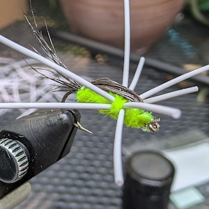 Op de afbeelding: Close-up van een vliegvislokaas. Het kunstaas heeft een felgroen lichaam met witte aanhangsels en donkerbruine en zilveren accenten. De haak wordt in een zwarte bankschroef gehouden, klaar om te worden gebonden.