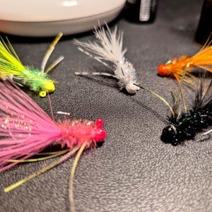 Op de afbeelding: Een verzameling kleurrijke vliegvisvliegen. De vliegen hebben verschillende kleuren, waaronder roze, groen, wit, oranje en zwart. Elke vlieg heeft een uniek ontwerp met veren en andere materialen, en kleine haken.