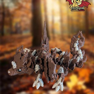May include: A brown articulated dragon figurine featuring textured scales and silver-colored accents on its claws, spine, and tail. The creature has a detailed head with a silver marking on its forehead and a logo reading Mythical Creature Rescue.