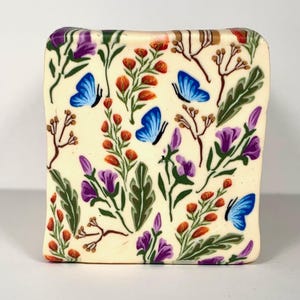 May include: A rectangular soap bar with a floral and butterfly design. The off-white bar features blue butterflies, purple flowers, orange buds, and green leaves. The soap has a slightly rounded edge.