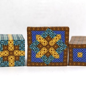 Puede incluir: Tres pequeños cubos de madera con intrincados patrones geométricos en tonos azules, amarillos y marrones. Los patrones son simétricos y crean un diseño visualmente atractivo.