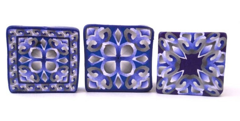 K&ouml;nnte beinhalten: Drei quadratische Keramikfliesen mit komplizierten geometrischen Mustern in Blau, Lila und Grau.