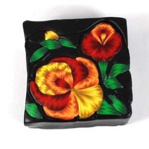 May include: A black square tile with three red, orange, and yellow pansies with green leaves.