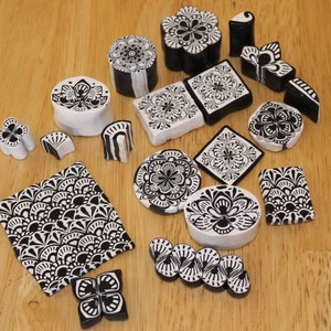 Può includere: Una collezione di pezzi decorativi in ceramica bianca e nera con motivi floreali e geometrici. I pezzi hanno varie forme, tra cui quadrati, cerchi e cilindri, e sono disposti su una superficie di legno. I disegni sono dettagliati e simmetrici.