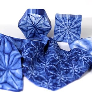 Puede incluir: Una tela azul con un patrón geométrico, doblada y mostrada con dos formas geométricas azules. La tela y las formas tienen un efecto tie-dye.