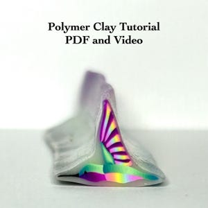 Puede incluir: Primer plano de una creación de arcilla polimérica con un diseño de arcoíris. La arcilla es una mezcla de colores, incluyendo morado, verde y amarillo. El texto "Polymer Clay Tutorial PDF and Video" está en la parte superior.