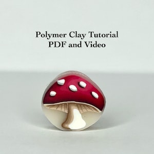 May include: A handcrafted polymer clay mushroom with a red cap and white spots. The mushroom has a beige stem and is set against a white background. Text at the top reads "Polymer Clay Tutorial PDF and Video."