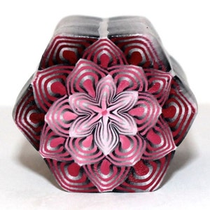 May include: A hexagonal polymer clay pendant with a pink and white flower design in the center. The flower is surrounded by concentric circles of red, white, and black.