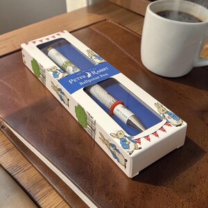 Puede incluir: Un bolígrafo Peter Rabbit en caja. La caja blanca presenta ilustraciones de Peter Rabbit y una pancarta azul con el texto "The World of Peter Rabbit Ballpoint Pen". El bolígrafo tiene una punta plateada y un anillo rojo.