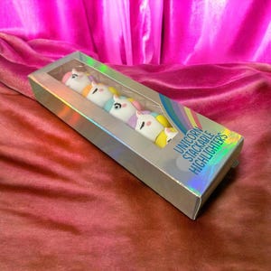 Puede incluir: Una caja rectangular plateada contiene cinco rotuladores de unicornio apilables. Cada rotulador es blanco con una melena y un cuerno de diferente color. La caja tiene el texto "UNICORN STACKABLE HIGHLIGHTERS" en azul.