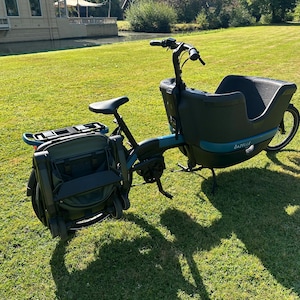 May include: A dark blue and black Gazelle cargo bike featuring a large front-mounted storage bin, a black seat, and a folded stroller attached to the rear rack. The bike has a sturdy frame, handlebars with grips, and black wheels on a grassy field.