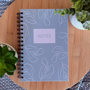 May include: A spiral-bound notebook with a light blue cover featuring white leaf illustrations. The word "NOTES" is printed in a rectangular lavender box. The notebook is on a wooden surface with small potted plants.