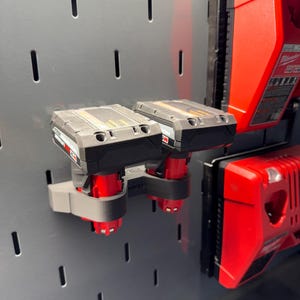 May include: Two black and red power tool batteries mounted on a gray pegboard wall with a red power tool charger on the right side of the image.