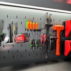May include: A gray pegboard wall with various tools and accessories hanging on it.  The tools include a red and black tape measure labeled "Milwaukee 25'" and a red and black hammer with a black handle.  There are also several orange and green screwdrivers and a black flashlight.