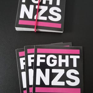 Könnte beinhalten: Ein Stapel quadratischer Aufkleber mit schwarzem Hintergrund, weißem Text FGHT NZS und horizontalen rosa Streifen oben und unten. Ein Stapel wird von einem dünnen roten Gummiband zusammengehalten, während ein weiterer Stapel darunter gefächert ist.