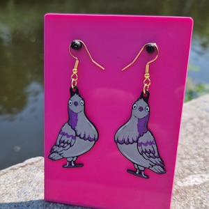 Puede incluir: Un par de pendientes colgantes con dibujos de palomas. Los pendientes son grises con detalles morados y contornos negros, colgados de ganchos dorados. Se exhiben sobre un fondo rosa brillante.