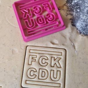 Könnte beinhalten: Ein rosa Kunststoff-Ausstecher mit dem Text "FCK CDU" wird in den Teig gedrückt und erzeugt einen Abdruck. Die Buchstaben sind deutlich sichtbar. Das Bild zeigt eine Nahaufnahme des Ausstechers und des Teigs.