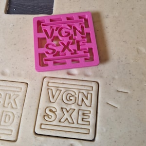 Könnte beinhalten: Ein rosa Ausstecher mit den Buchstaben "VGN SXE" liegt auf einem Keks mit den gleichen Buchstaben. Der Ausstecher ist quadratisch mit einem gitterartigen Design. Der Keks ist hellbraun.
