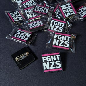 Può includere: Una collezione di toppe rettangolari bianche e nere con le scritte "FGHT NZS" e "SZN FGHT" in lettere bianche su una striscia rosa. Alcune toppe sono confezionate singolarmente in plastica trasparente. Una toppa è attaccata a un quadrato nero con una spilla da balia.