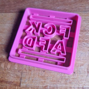 Könnte beinhalten: Ein leuchtend pinkfarbener Ausstecher mit den Worten "A F*CK" in Großbuchstaben. Die Buchstaben sind fett und ohne Serifen. Der Ausstecher ist rechteckig mit einem Gittermuster, ideal zum Backen.